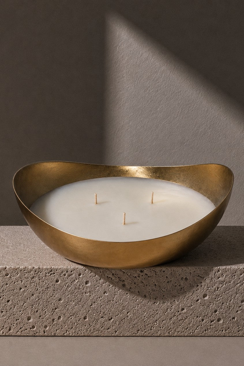 A three-wick soy candle in a polished gold-toned metal bowl, displayed on a textured gray stone block against a dark gray background with dramatic diagonal lighting.