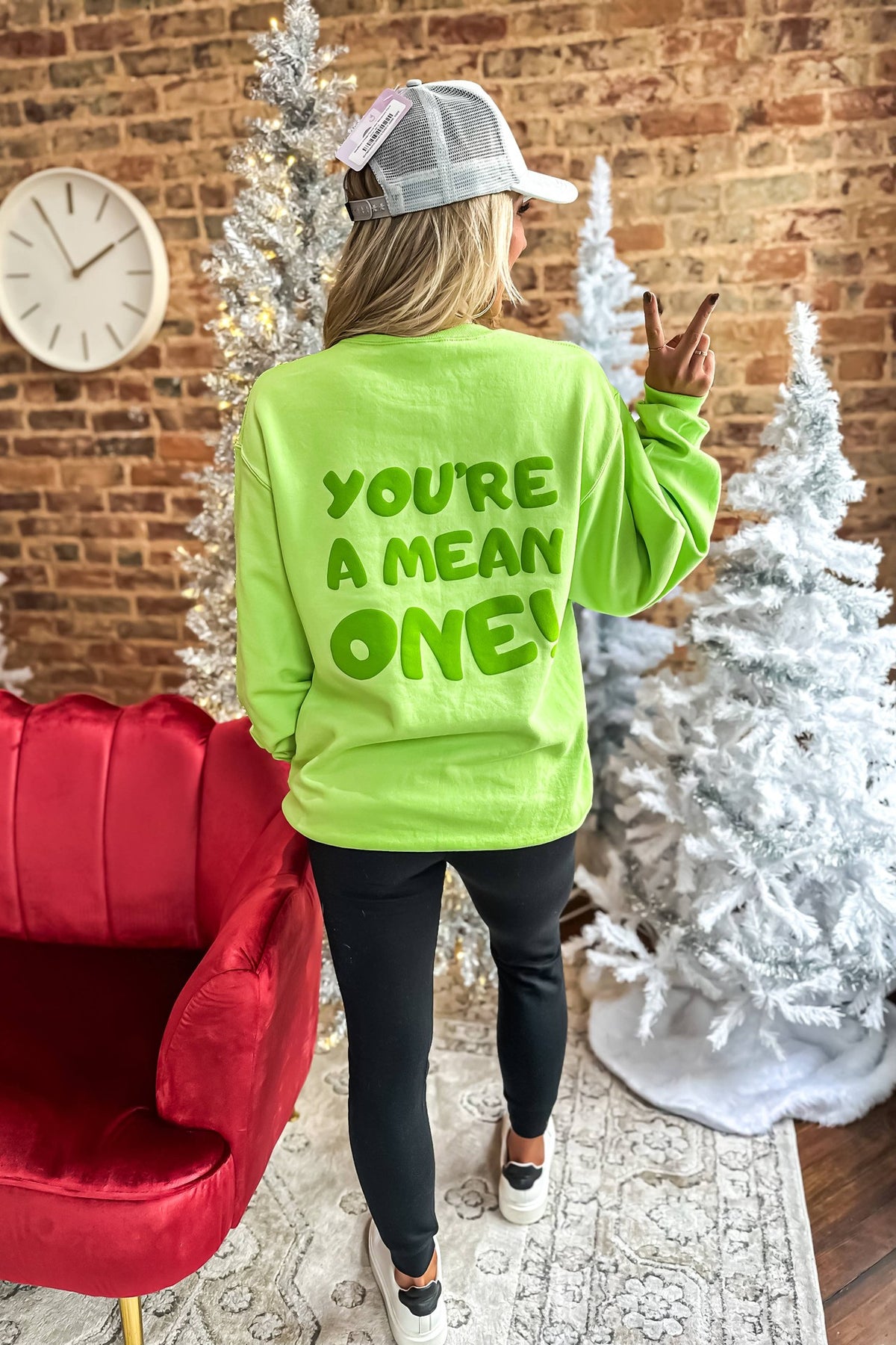 A person wearing a green crewneck sweatshirt with the text 'YOU'RE A MEAN ONE!' printed on the front, standing in a room with Christmas decorations.