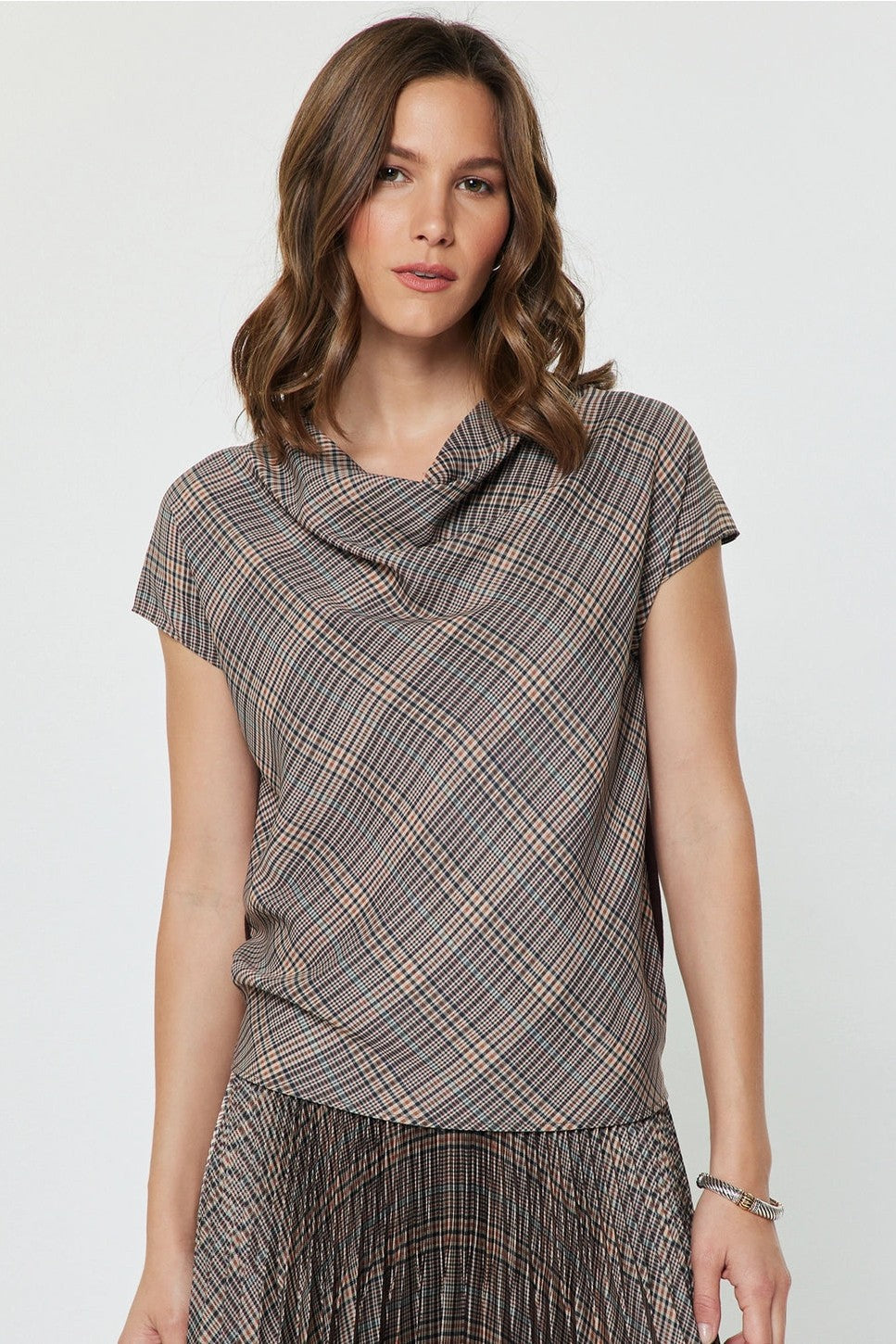 A woman wearing a short-sleeve cowl-neck top with a brown, gray, and beige plaid pattern, paired with a matching pleated skirt, against a plain light gray background.