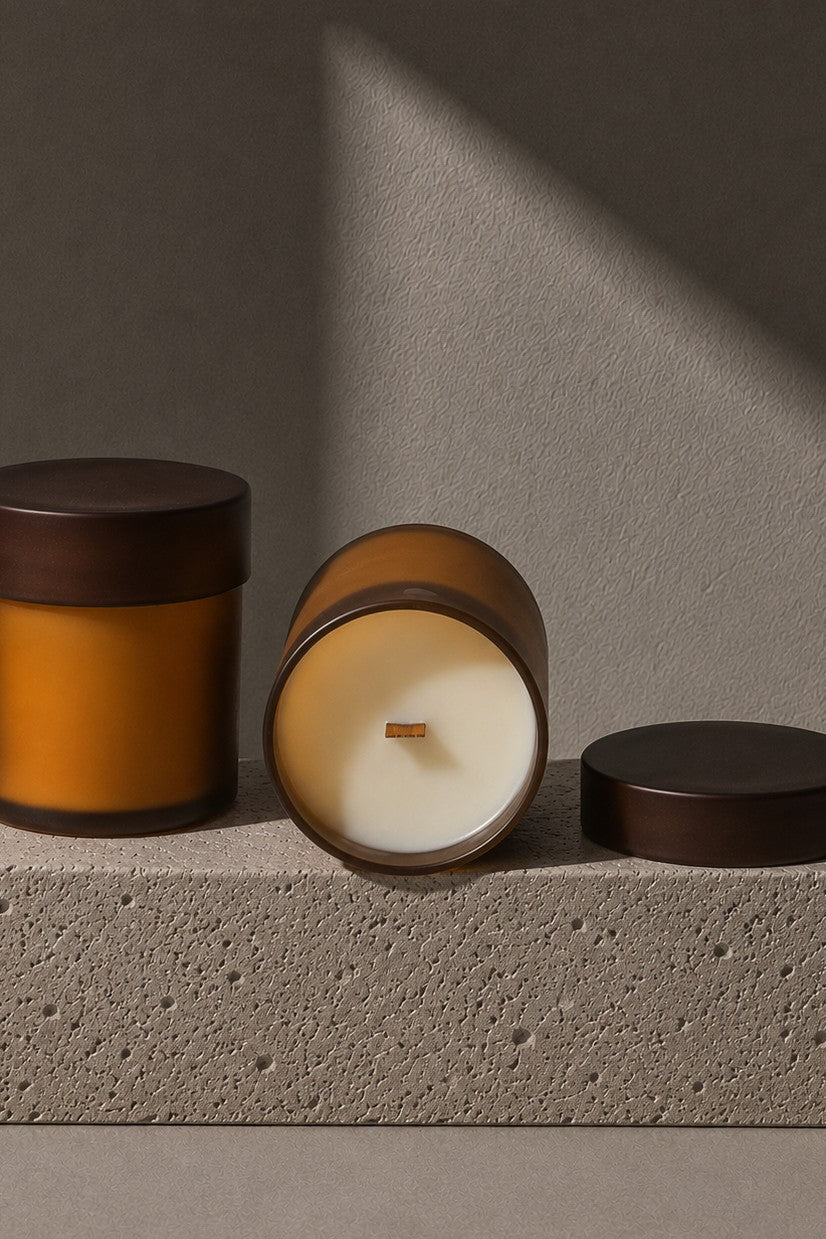 Three amber glass jar candles with dark brown lids are arranged on a textured stone block against a warm gray background with dramatic diagonal sunlight and shadows.