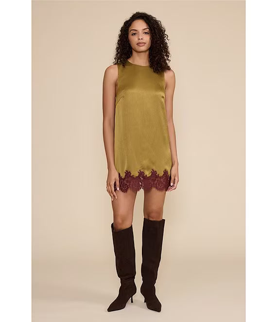 A woman modeling a sleeveless olive green satin mini dress with a burgundy lace hem, paired with dark brown knee-high boots, against a solid beige studio background.