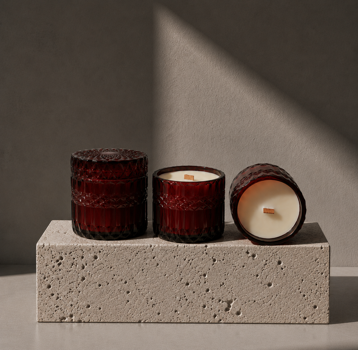 Three amber glass jar candles arranged on a textured stone block against a gray wall with dramatic diagonal shadows. One candle is upright and closed, one is upright and open showing cream wax, and one is lying on its side showing the wax surface and wick.