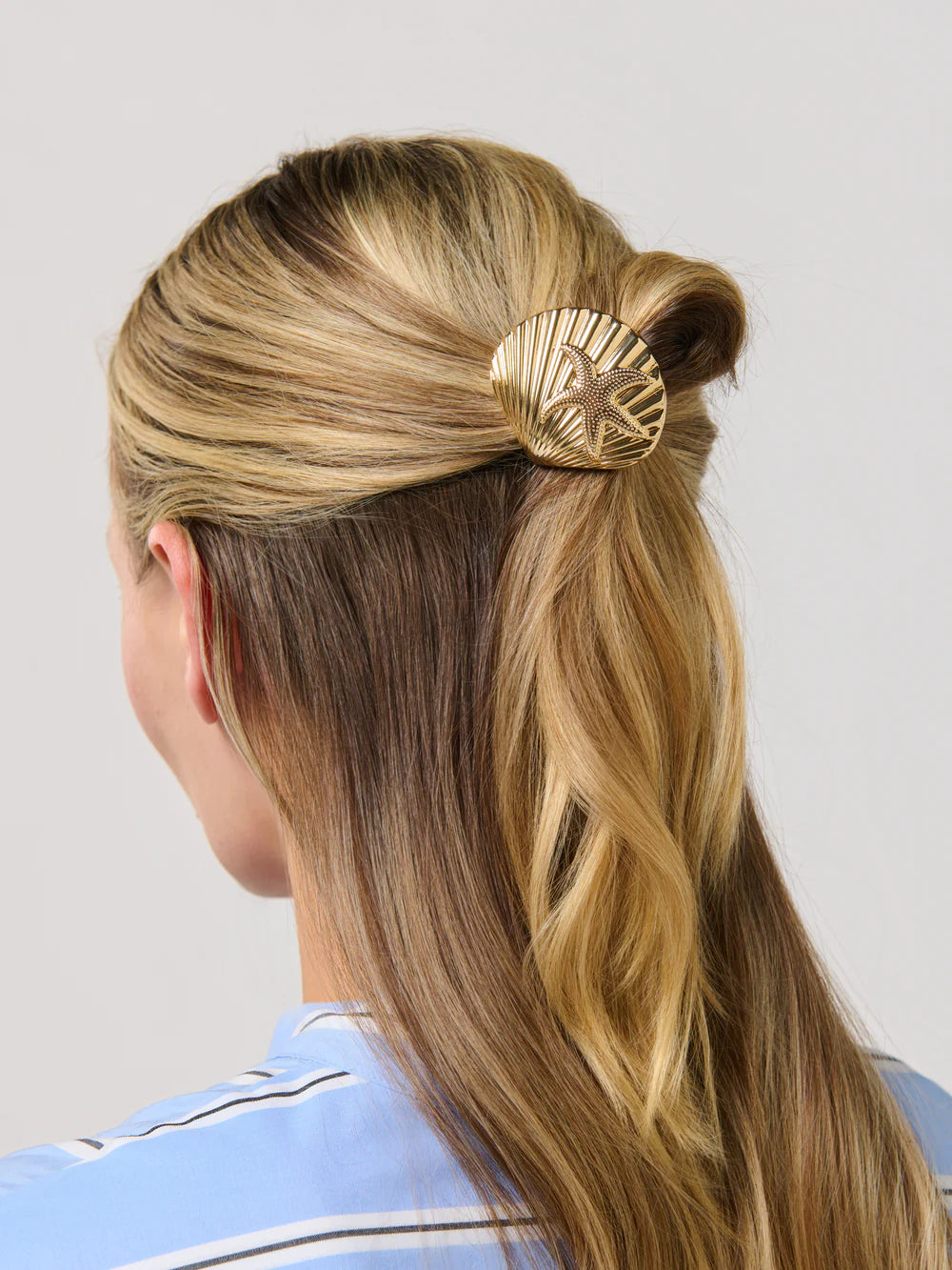Woman with blonde hair styled in a ponytail with a gold hair accessory against a light gray background