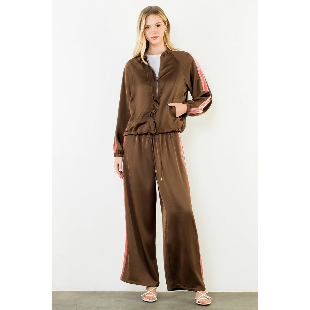A woman modeling a brown satin two-piece tracksuit with pink stripe details on the sleeves and legs, standing against a plain off-white wall.