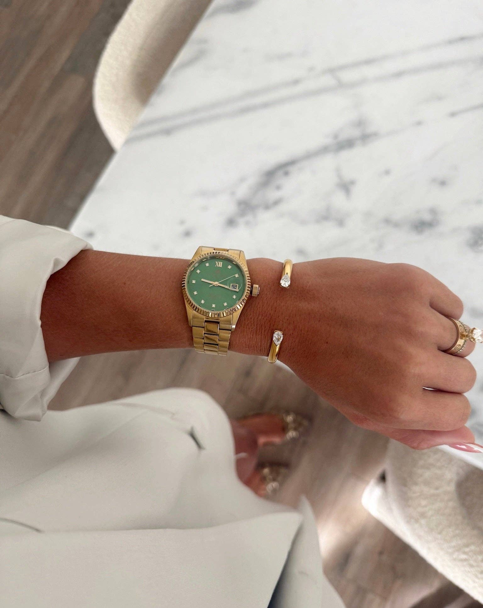 A gold-tone watch with a green dial and bezel is worn on a person's wrist, alongside matching gold rings and a bracelet, in a lifestyle setting with a marble tabletop and wooden floor in the background.