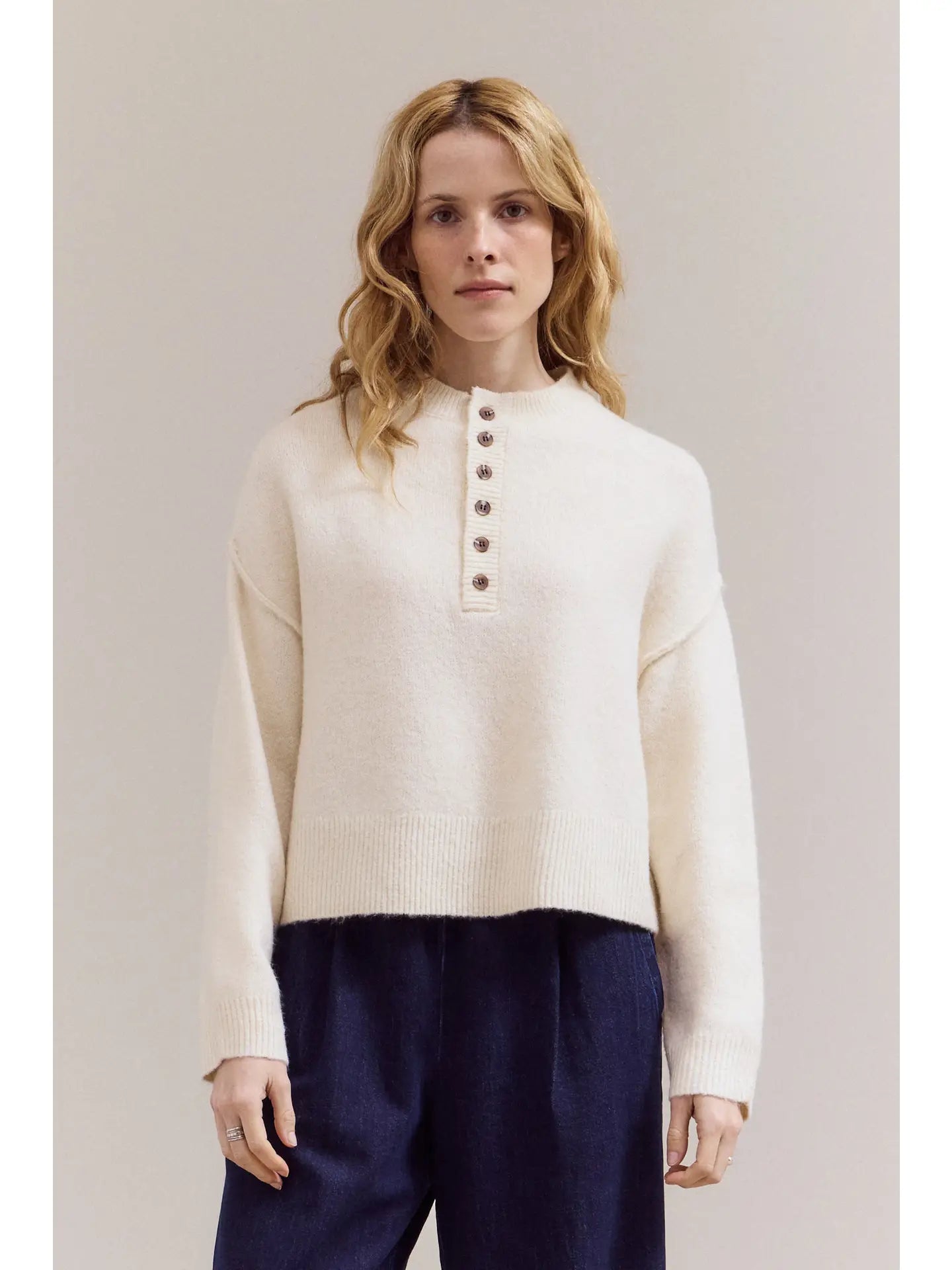 Woman wearing a cream-colored sweater with buttons and dark pants against a plain background