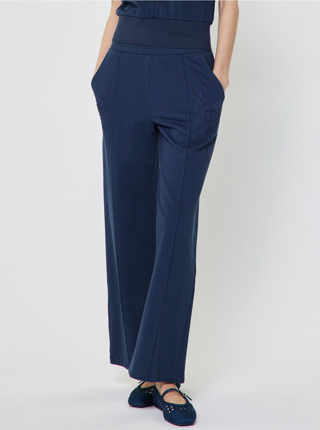 A person wearing navy blue wide-leg trousers with side pockets and matching navy top, paired with dark blue flat shoes, photographed against a light gray studio background.