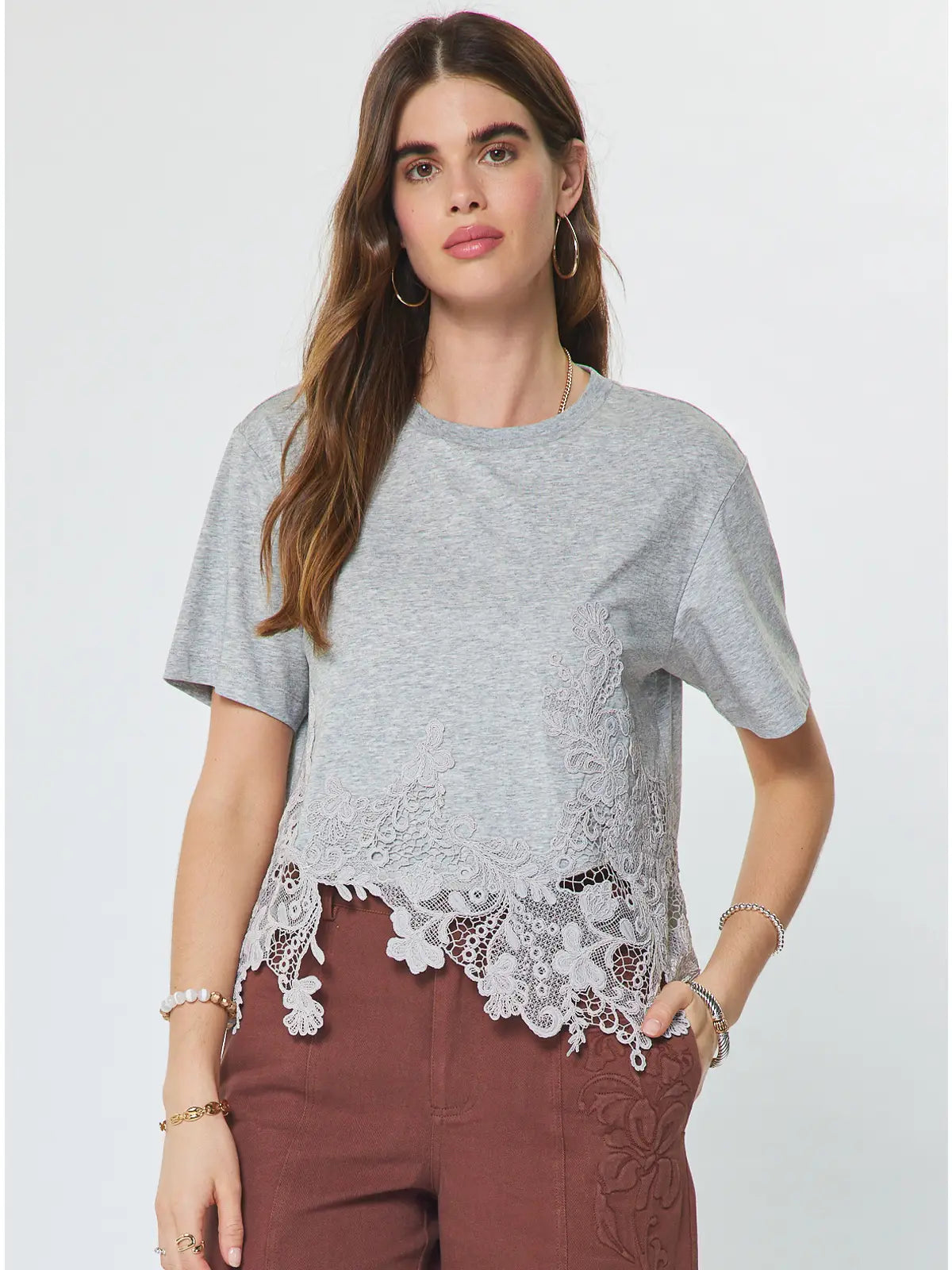 A woman modeling a light gray short-sleeve t-shirt with white floral lace trim along the hem, paired with brown pants, against a plain light gray background.