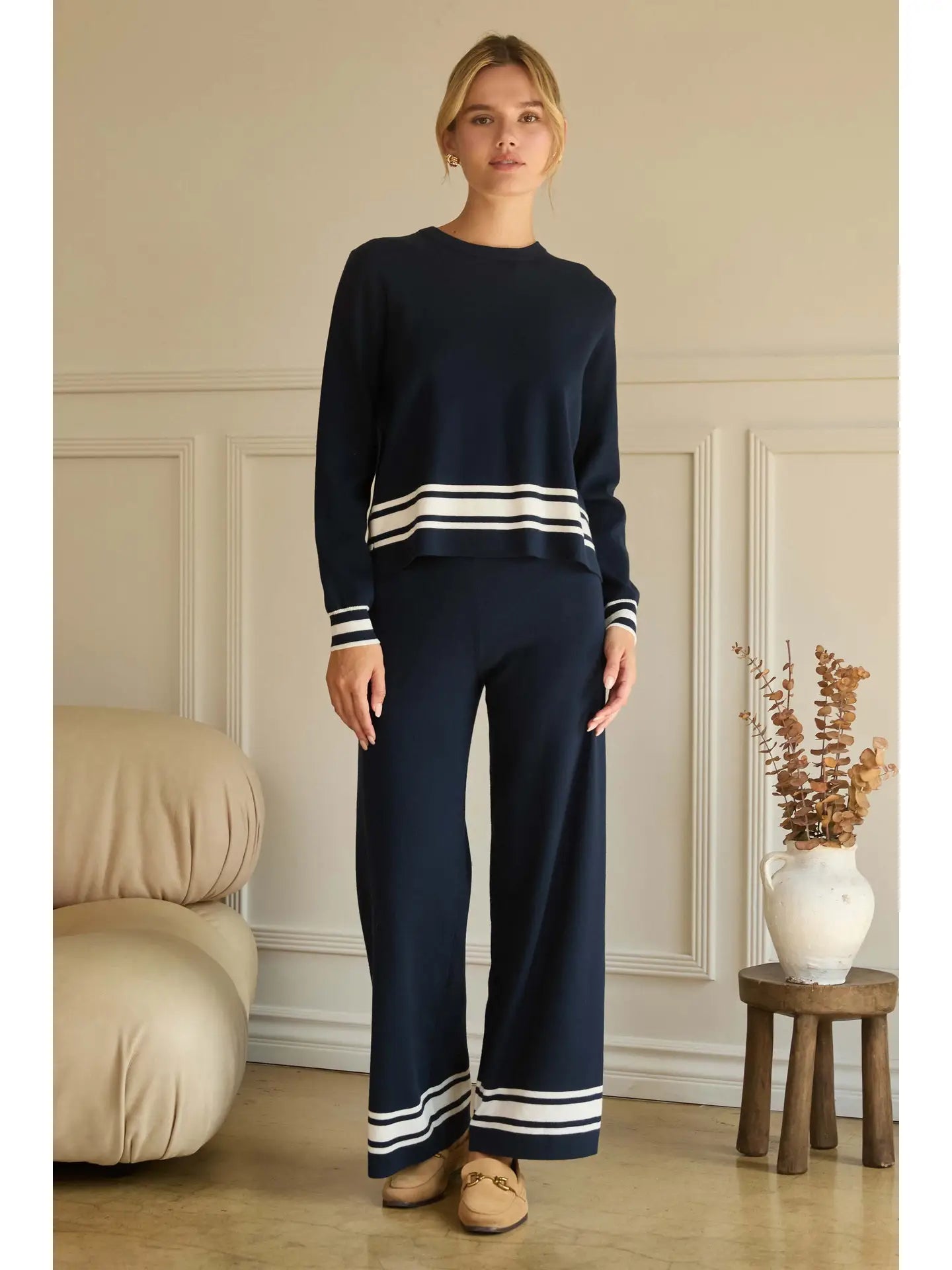 Woman wearing a navy sweater and pants with white stripes in a room with beige walls and wooden floor.