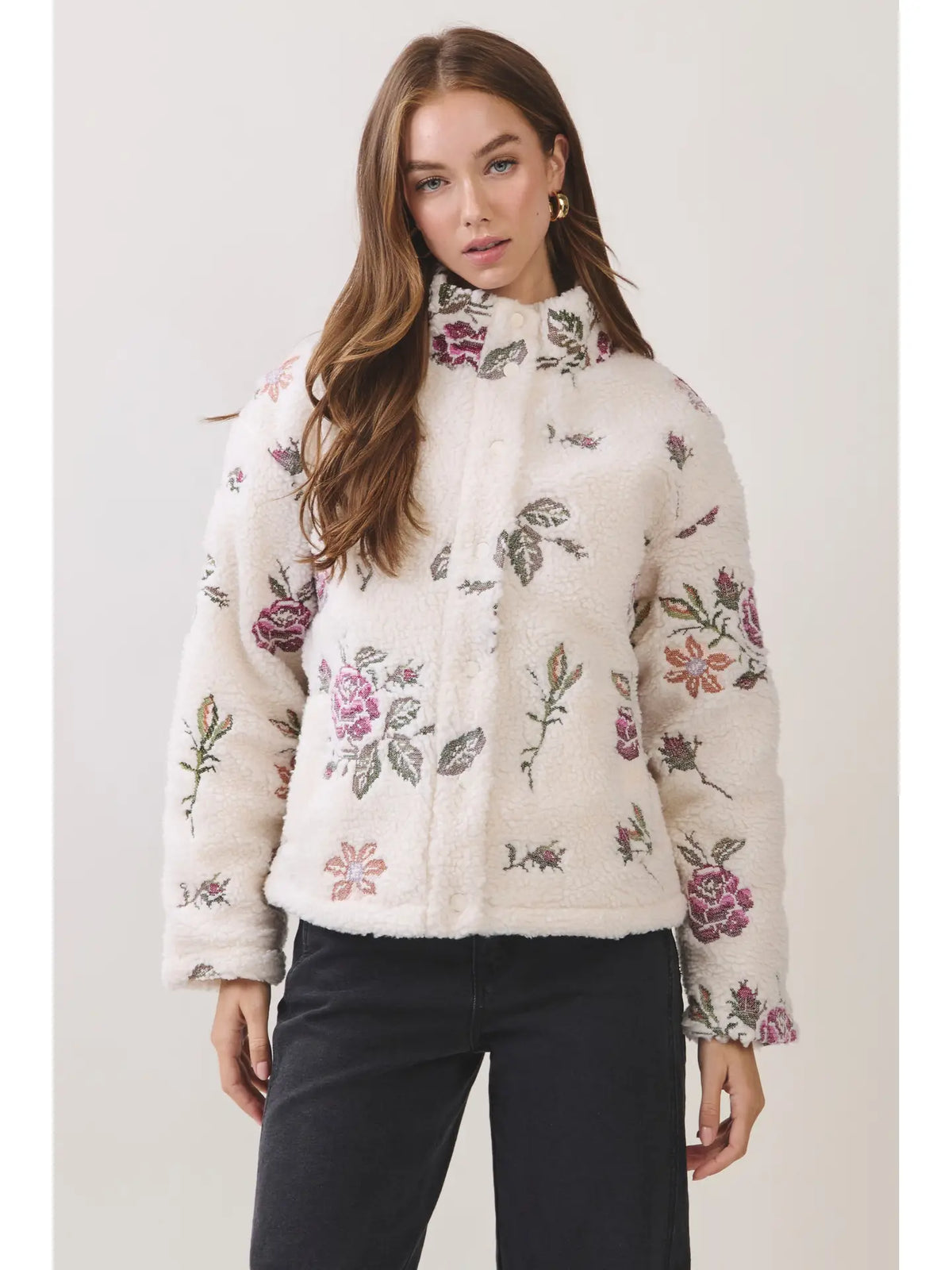 A woman modeling a cream-colored sherpa jacket with a pink and green floral pattern, paired with black pants, against a light gray studio background.