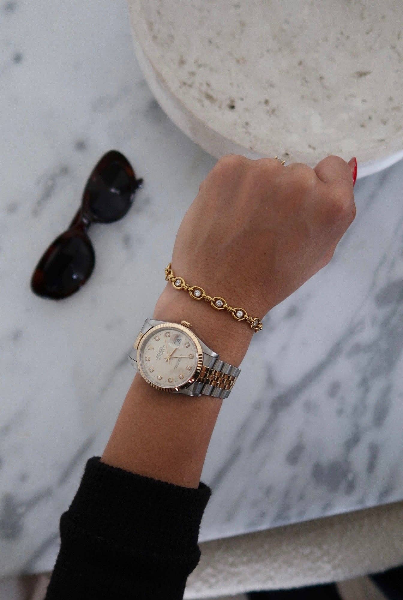 Hand wearing a watch and bracelet on a marble surface with sunglasses.