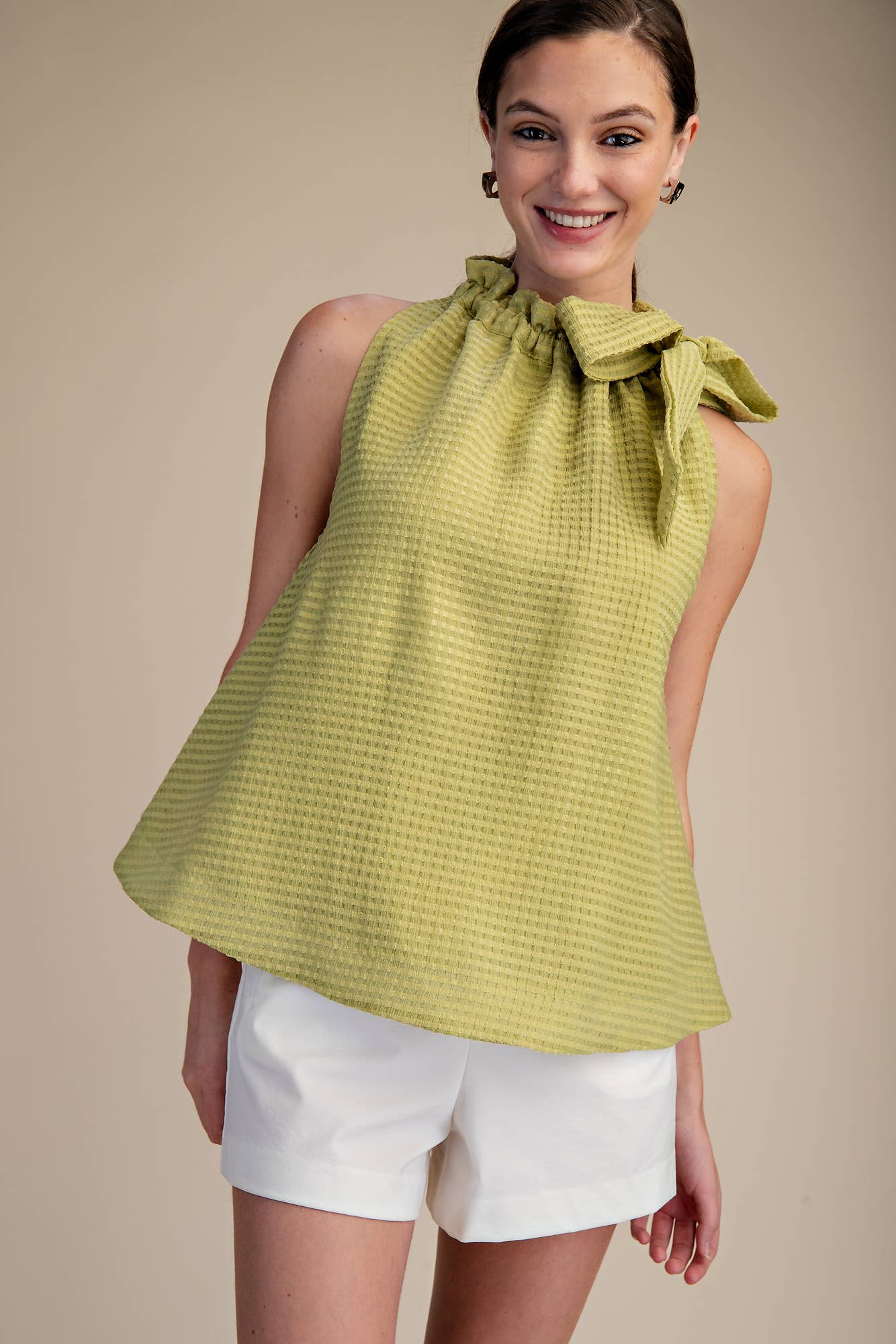Woman wearing a green sleeveless top with a bow neckline and white shorts on a beige background