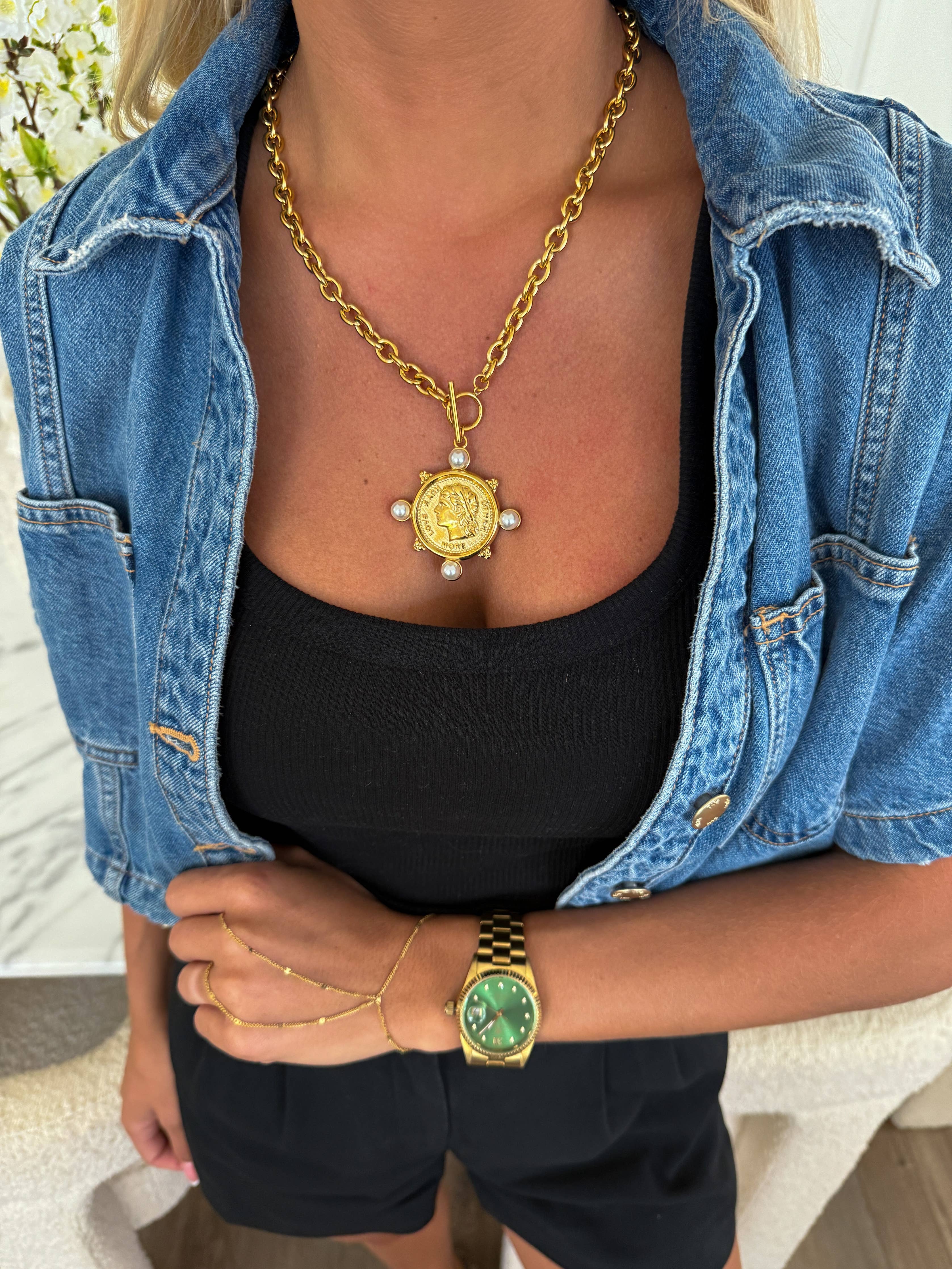 Person wearing a gold necklace and green watch with a denim jacket and black top.