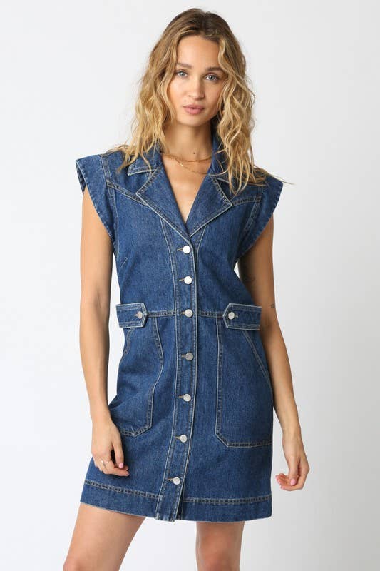 A woman modeling a sleeveless blue denim dress with a button-front closure and front patch pockets, photographed against a plain light gray studio background.