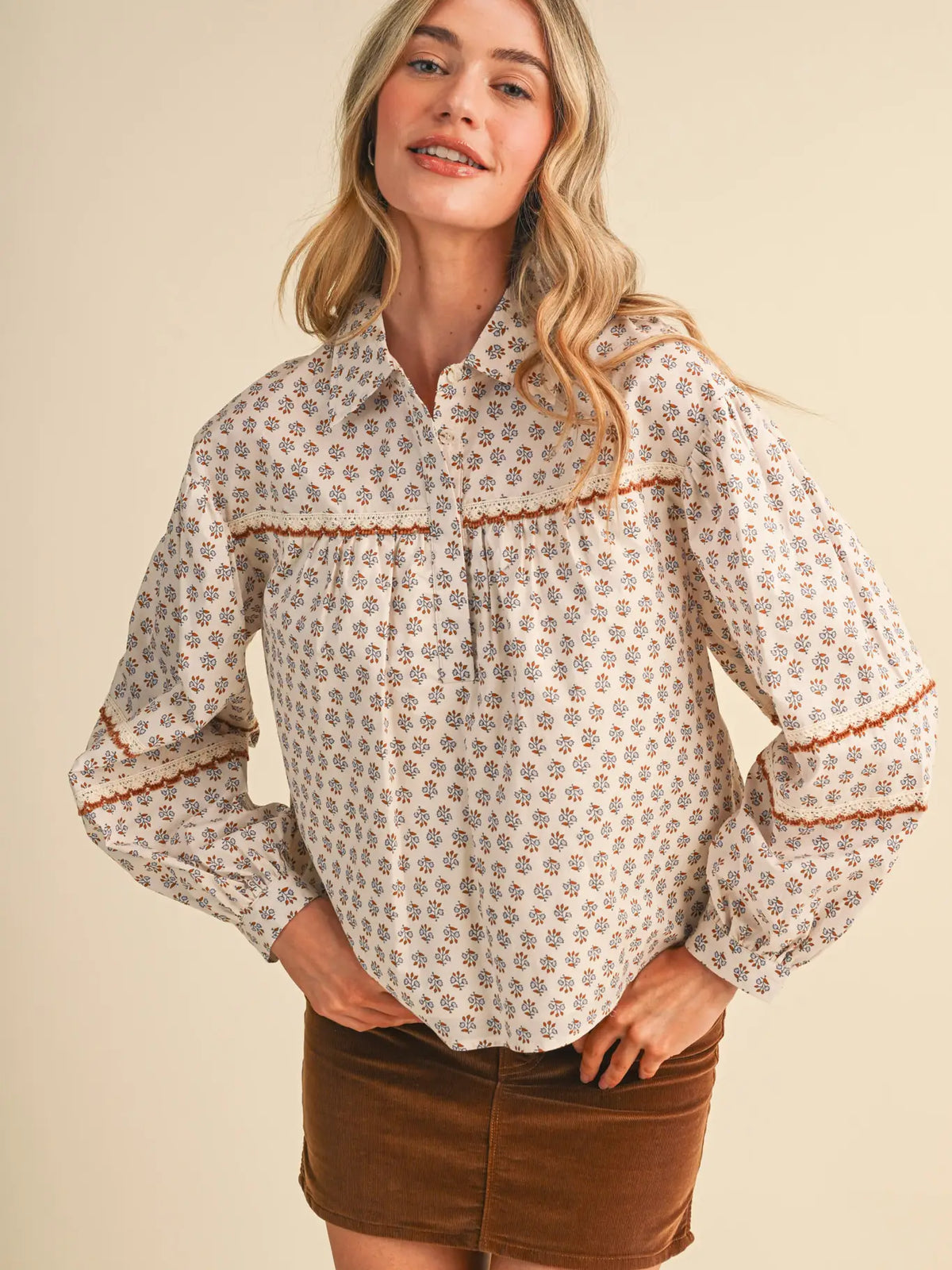 A woman wearing a cream-colored long-sleeve blouse with a small brown and beige floral pattern, brown ruffle trim, and a brown corduroy mini skirt, posing against a solid beige background.