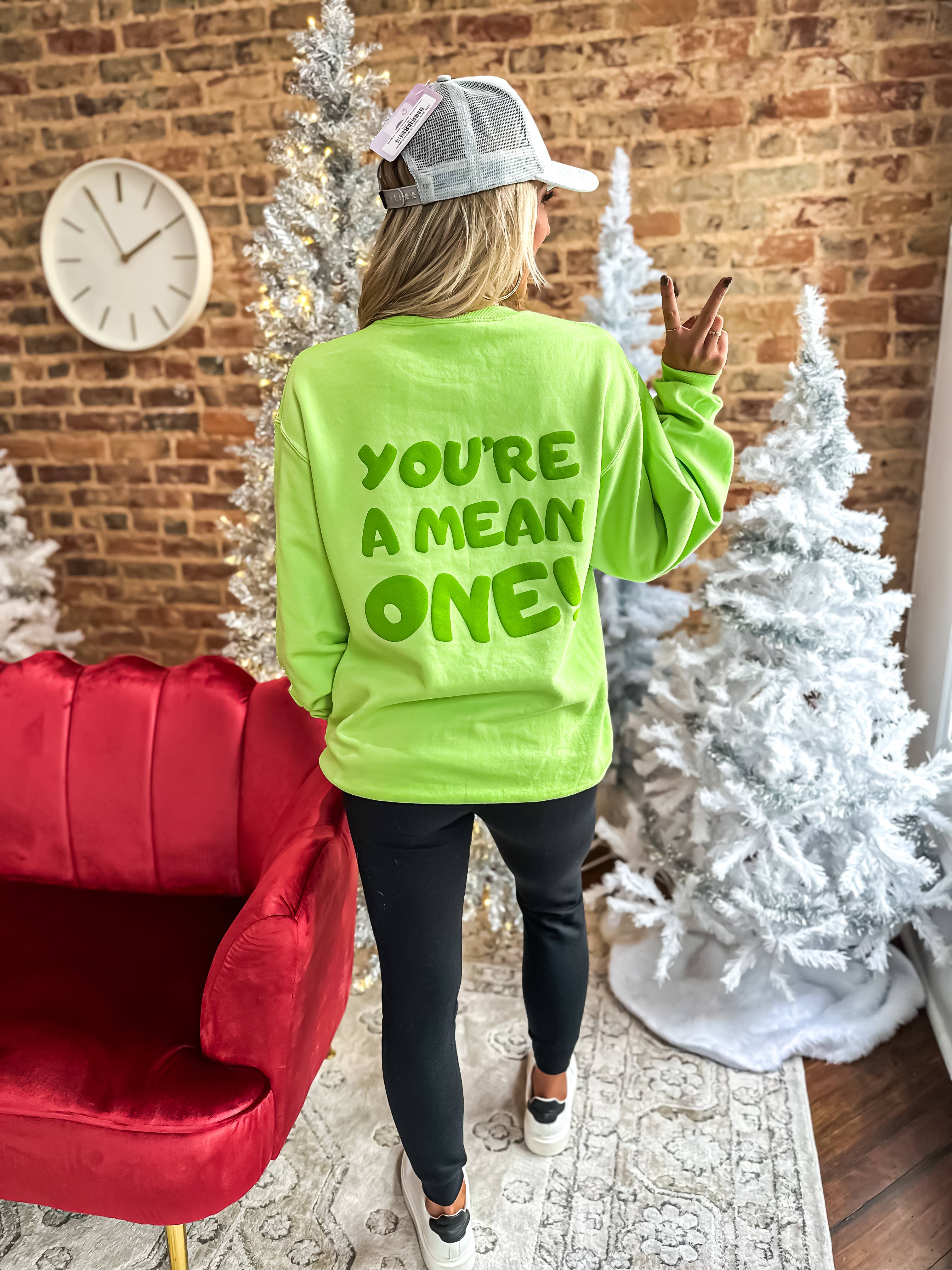 A person wearing a green crewneck sweatshirt with the text 'YOU'RE A MEAN ONE!' printed on the front, standing in a room with Christmas decorations.