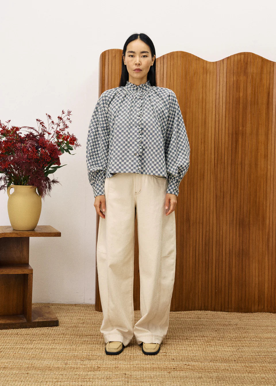 Woman wearing a checkered blouse and beige pants standing in front of a wooden screen with a vase of flowers on a table.