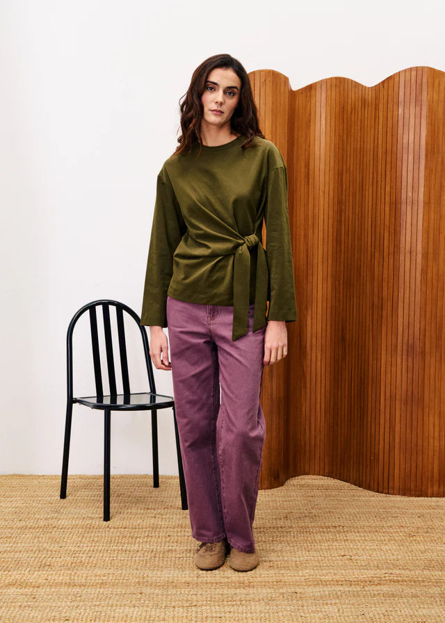 Woman wearing a green top and purple pants standing in front of a wooden screen.