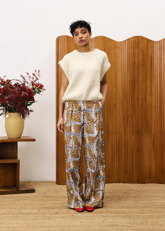 Woman wearing a white sweater and patterned pants standing in front of a wooden screen with a vase of flowers on a table.