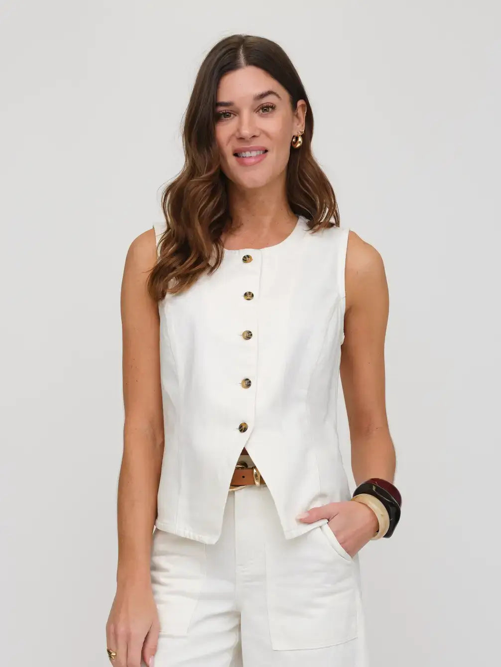 Woman wearing a white sleeveless button-up top with gold buttons on a plain background