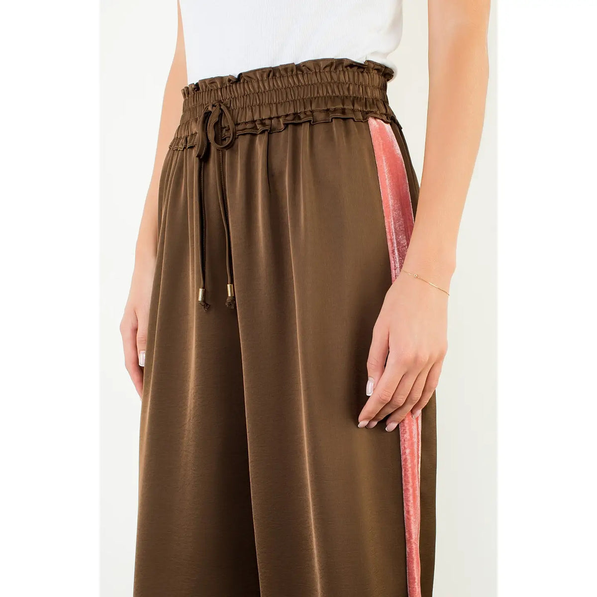 A close-up of a model wearing brown wide-leg satin pants with a pink textured side stripe and a matching brown drawstring waist.