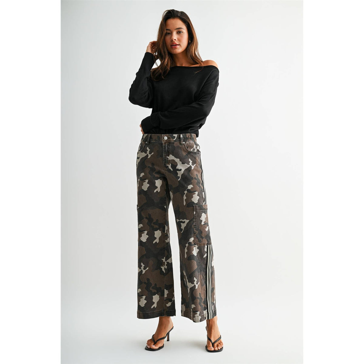 A woman modeling a pair of wide-leg cropped pants with a brown, gray, and beige camouflage pattern, paired with a black off-the-shoulder long-sleeve top and black heeled sandals, against a light gray studio background.