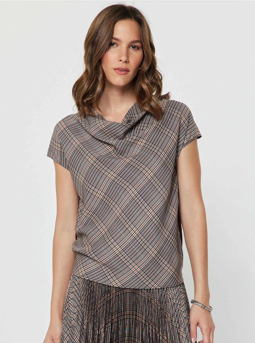 A woman wearing a short-sleeve cowl-neck top with a brown, gray, and beige plaid pattern, paired with a matching pleated skirt, against a plain light gray background.