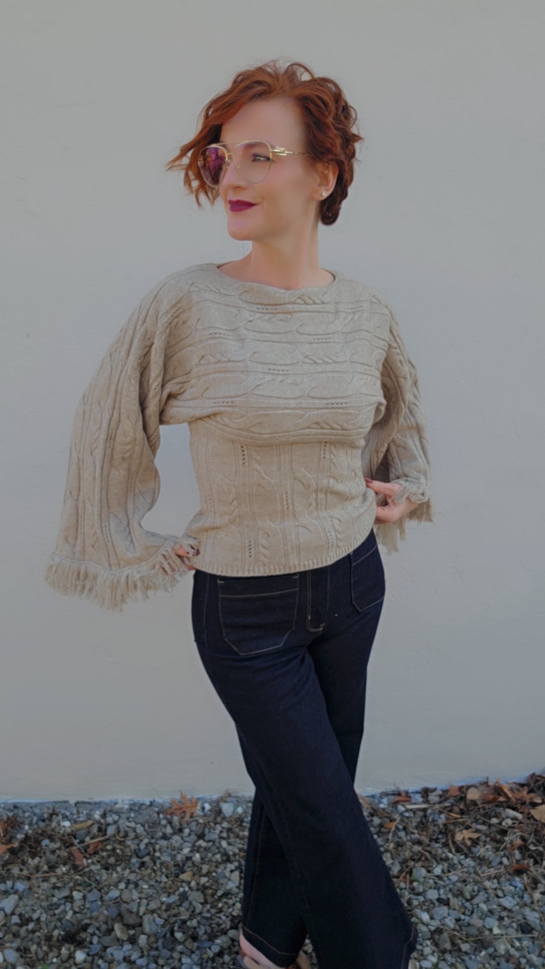 Woman wearing a beige textured top with long sleeves and dark jeans against a neutral background.