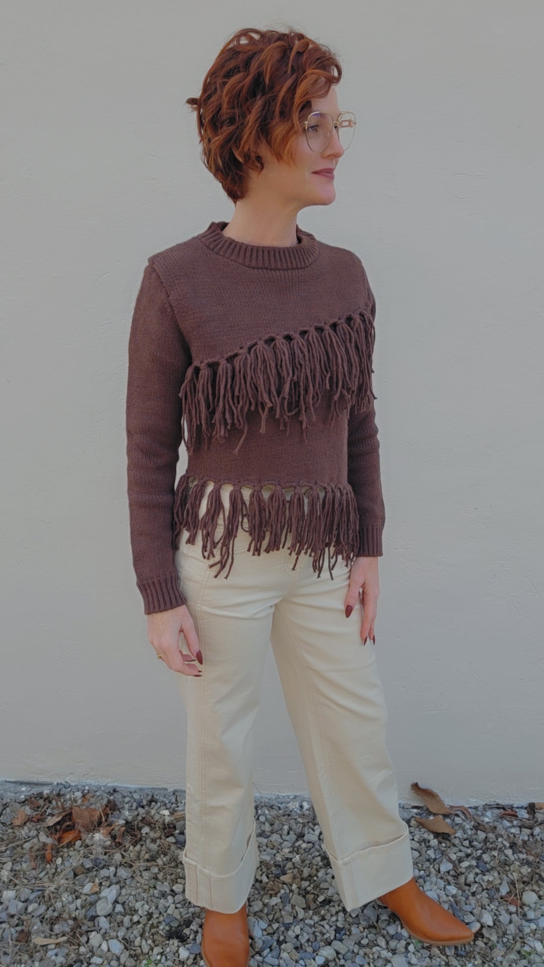 Woman wearing a brown fringed sweater and beige pants against a plain background