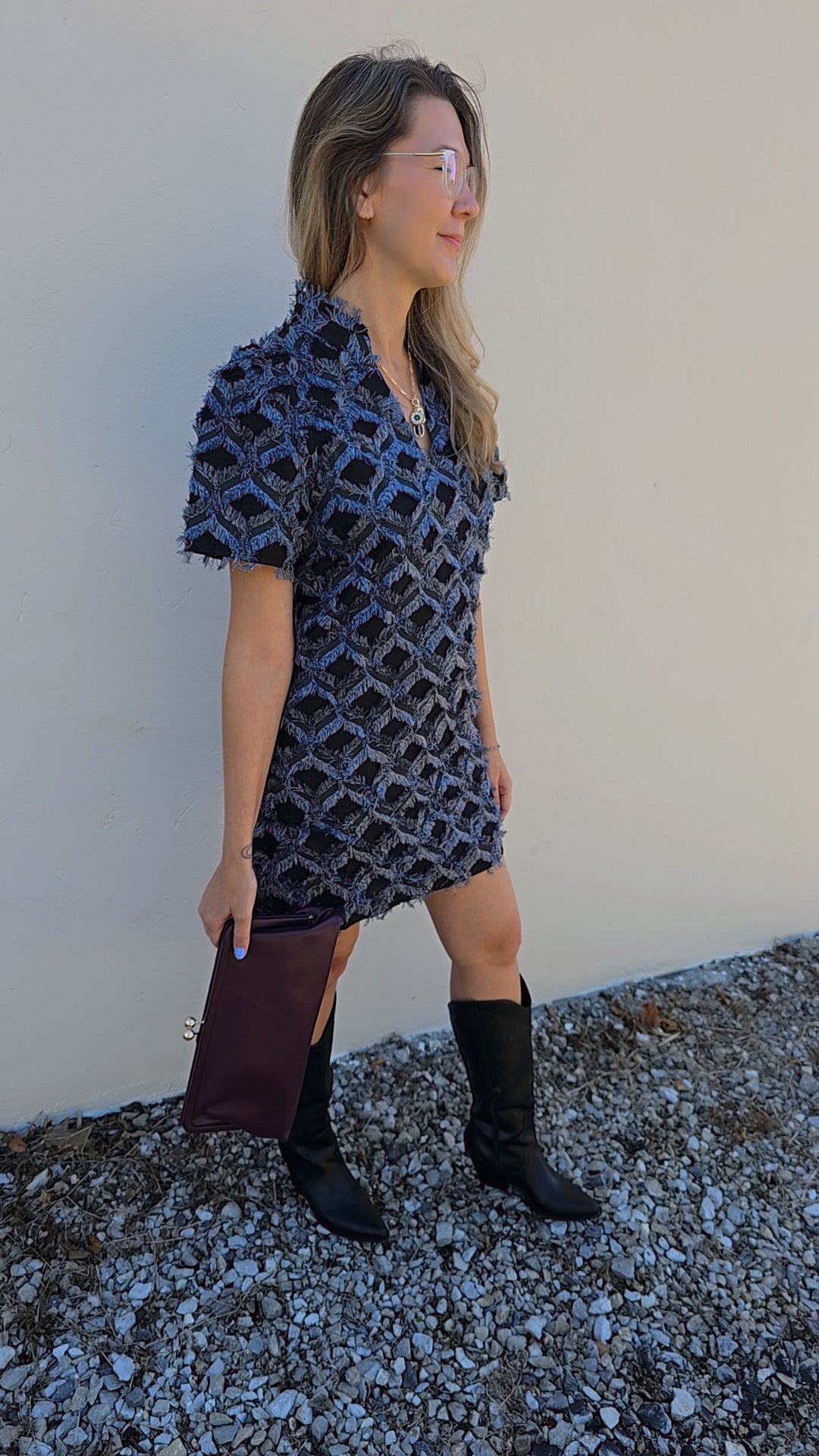 Woman wearing a patterned dress and boots standing against a plain wall.