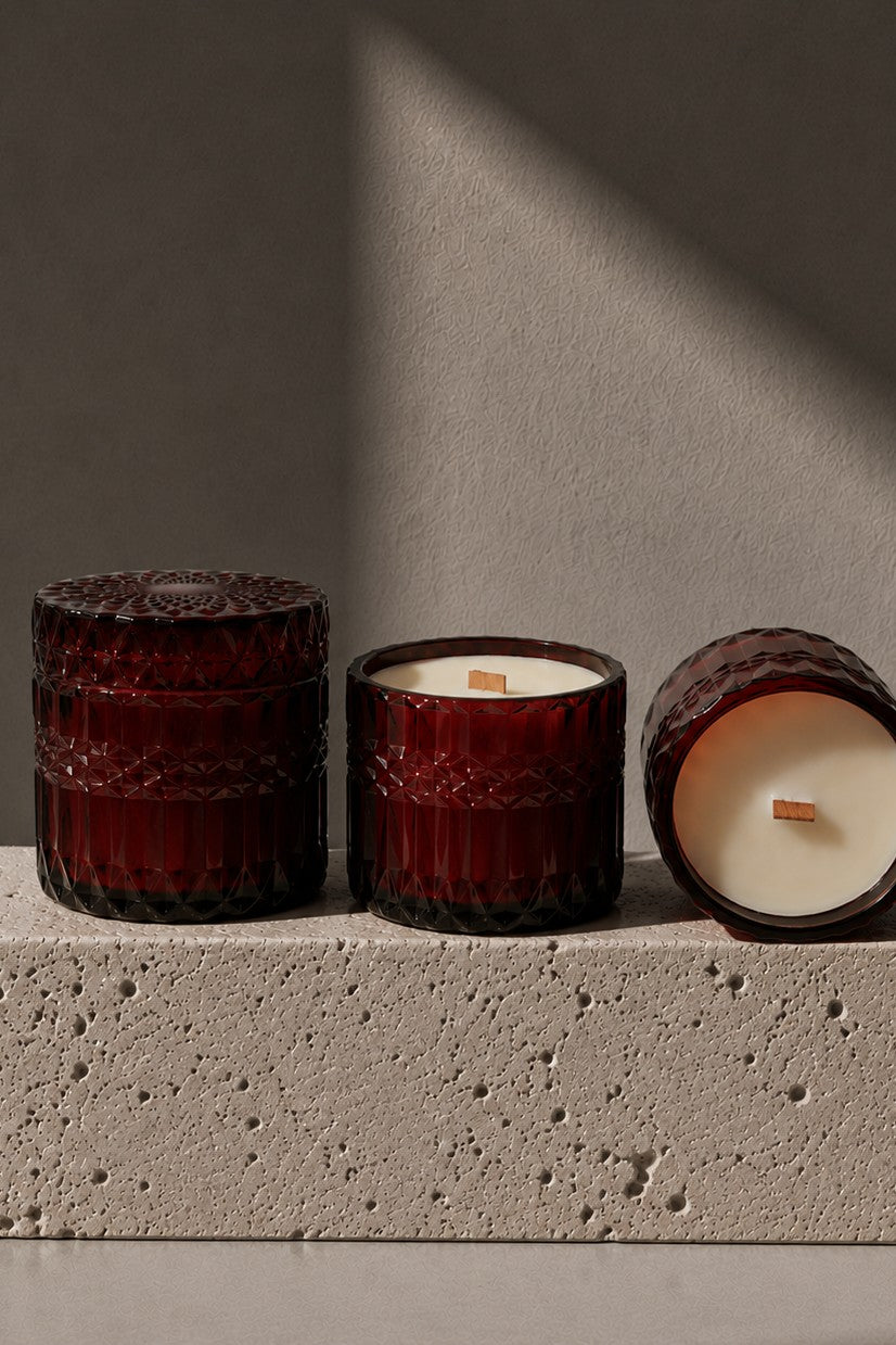 Three amber glass jar candles arranged on a textured stone block against a gray wall with dramatic diagonal shadows. One candle is upright and closed, one is upright and open showing cream wax, and one is lying on its side showing the wax surface and wick.