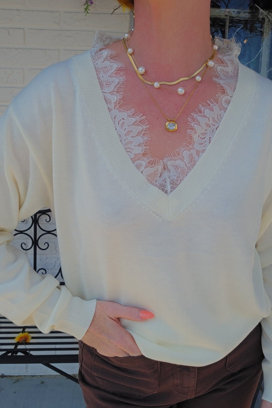 A person wearing a cream-colored knit V-neck sweater over a sheer white lace camisole, accessorized with layered gold necklaces, paired with brown pants.