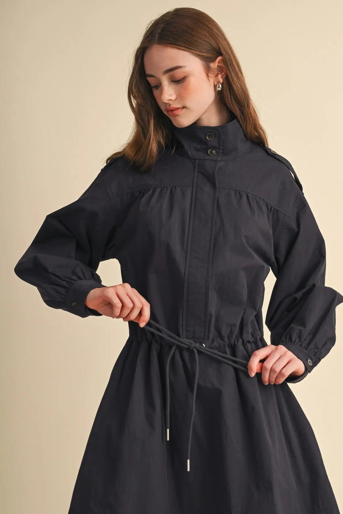 A woman wearing a dark charcoal gray high-neck long-sleeve dress with a drawstring waist, standing against a solid beige background.
