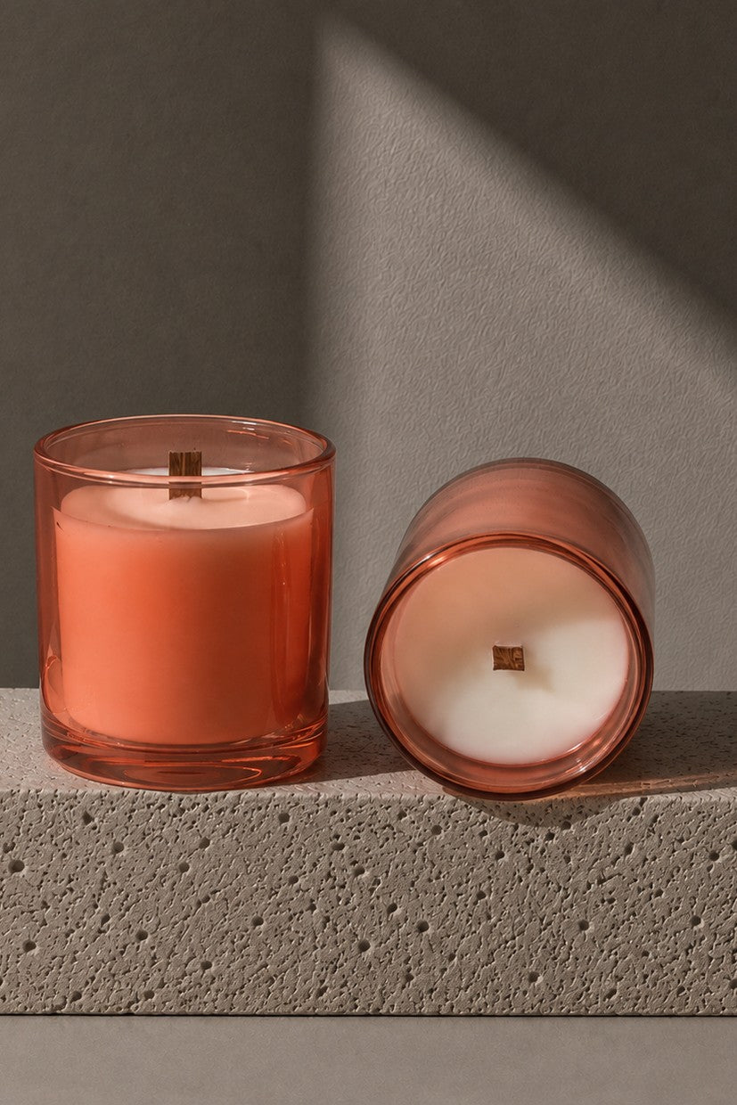 Two amber glass jar candles on a textured gray stone block, one standing upright and one lying on its side to show the white wax surface, against a warm gray background with dramatic diagonal shadows.