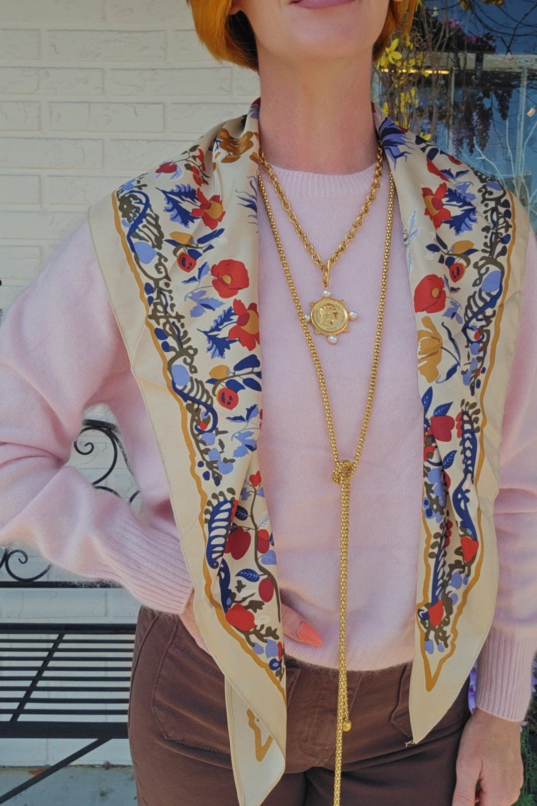 A woman wearing a light pink sweater and brown pants, styled with a cream-colored silk scarf featuring a colorful floral and botanical print, layered gold necklaces, and a red bob haircut.