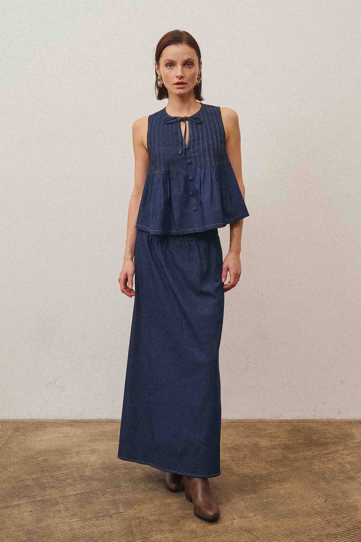 Woman wearing a navy blue sleeveless top and long skirt standing against a plain wall.