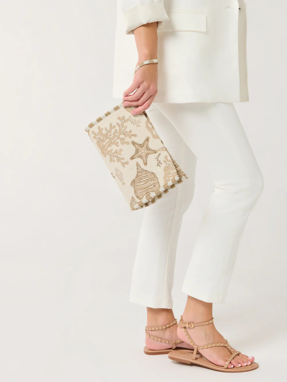 Person holding a decorative clutch with white pants and sandals on a light background