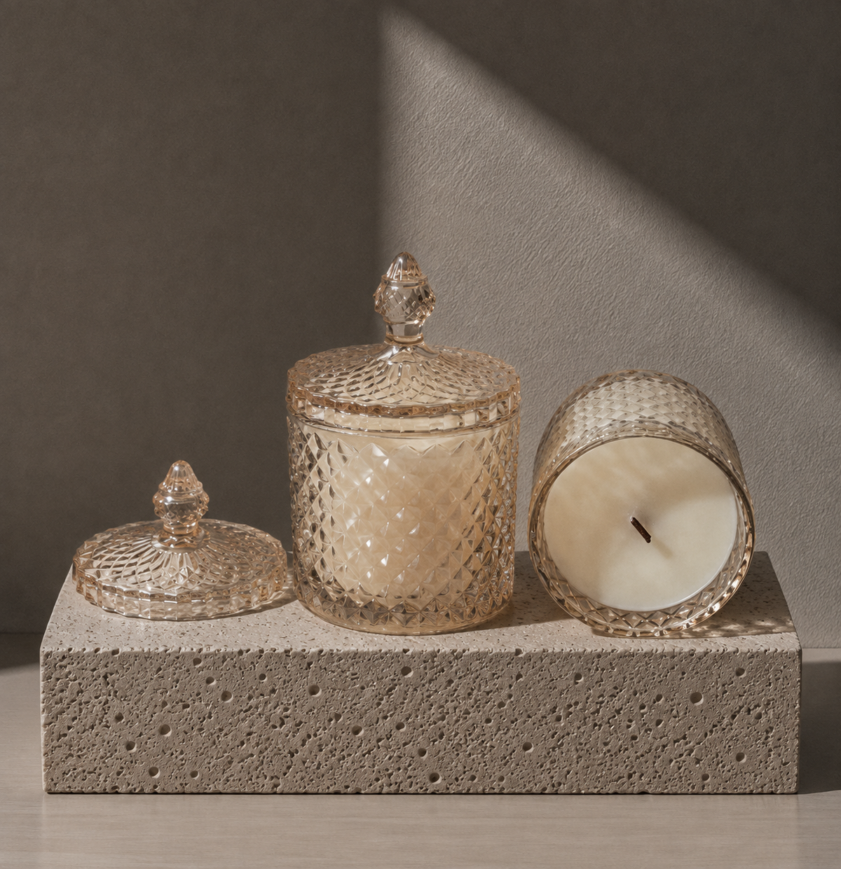 Three textured glass candle containers with matching lids are arranged on a rough stone block against a warm gray wall. One container is upright and closed, one is tipped on its side showing the cream wax inside, and one lid rests beside the upright piece.