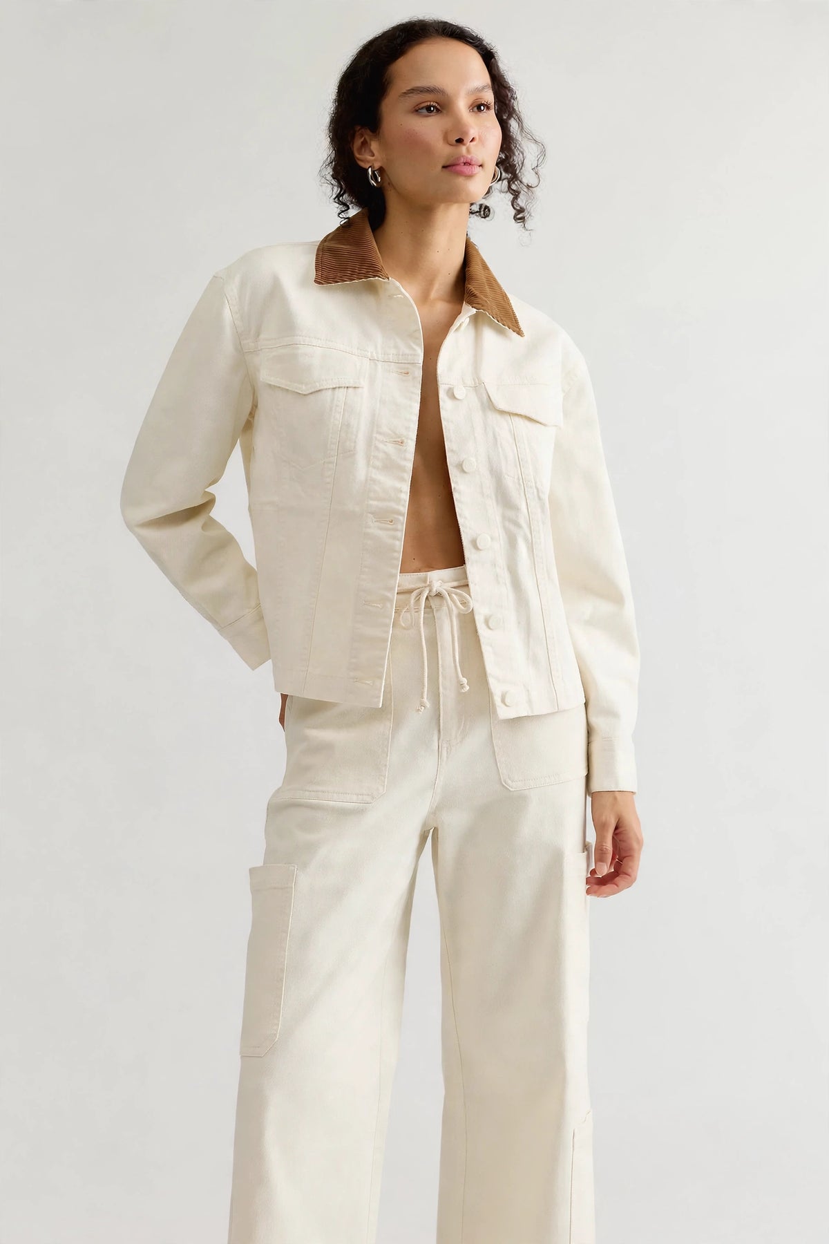 A model wearing a cream-colored denim jacket with a brown corduroy collar and matching cream wide-leg trousers, standing against a light gray studio background.