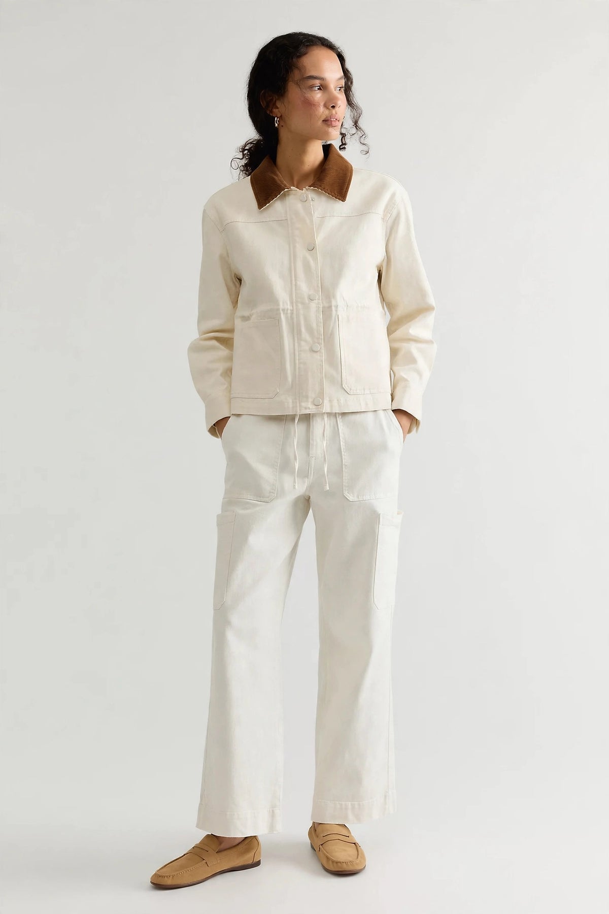 A woman modeling a cream-colored jacket with a contrasting brown collar, paired with matching cream wide-leg trousers and tan loafers, against a plain light gray studio background.