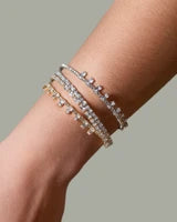 Three slim diamond tennis bracelets in silver, yellow gold, and rose gold tones stacked on a person's wrist, set against a solid gray background.