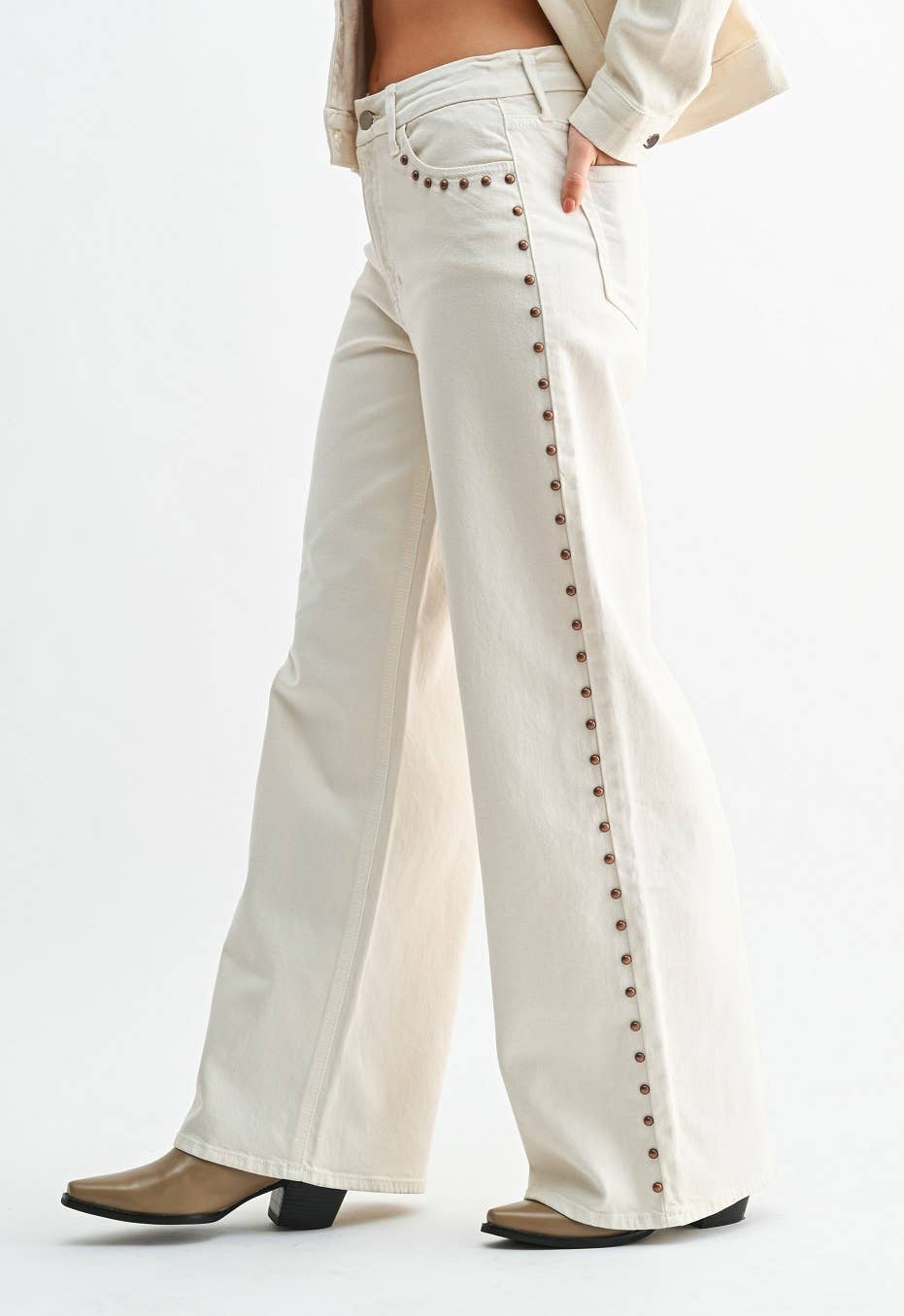 A person wearing off-white wide-leg jeans with a vertical row of dark brown stud embellishments down the outer side, paired with tan heeled boots against a plain white studio background.