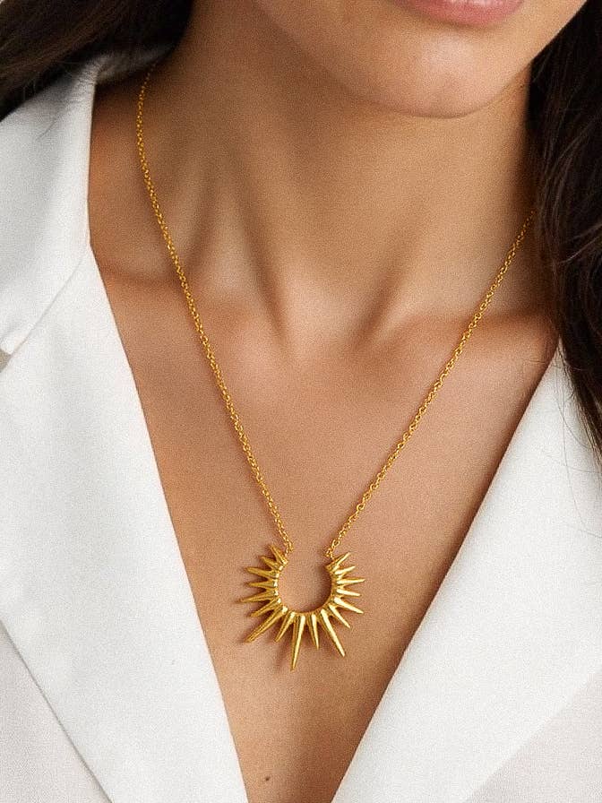 Gold necklace with a sunburst pendant worn by a person in a white outfit.