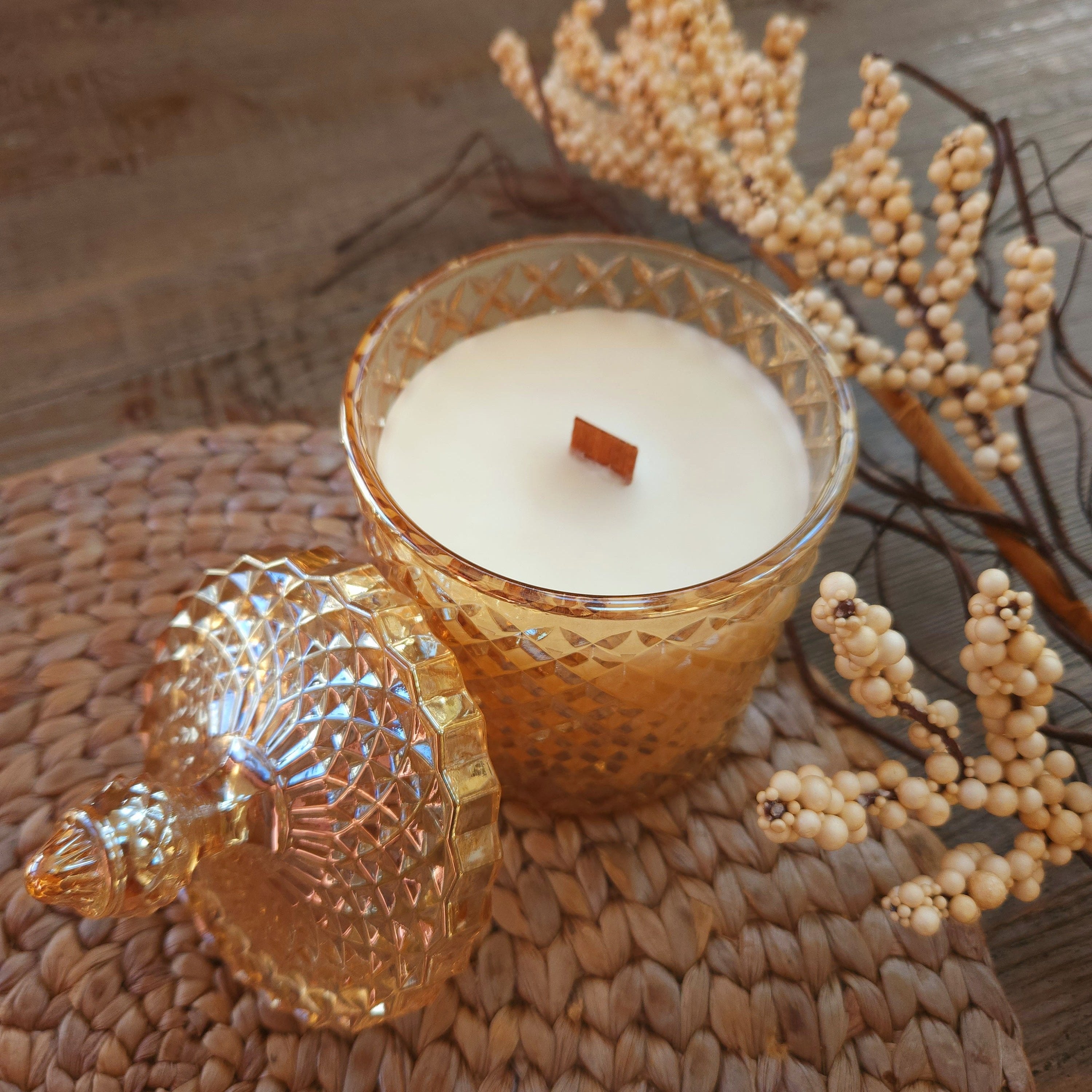 Decorative candle in a textured glass holder with a shell design, surrounded by dried plants on a woven surface.