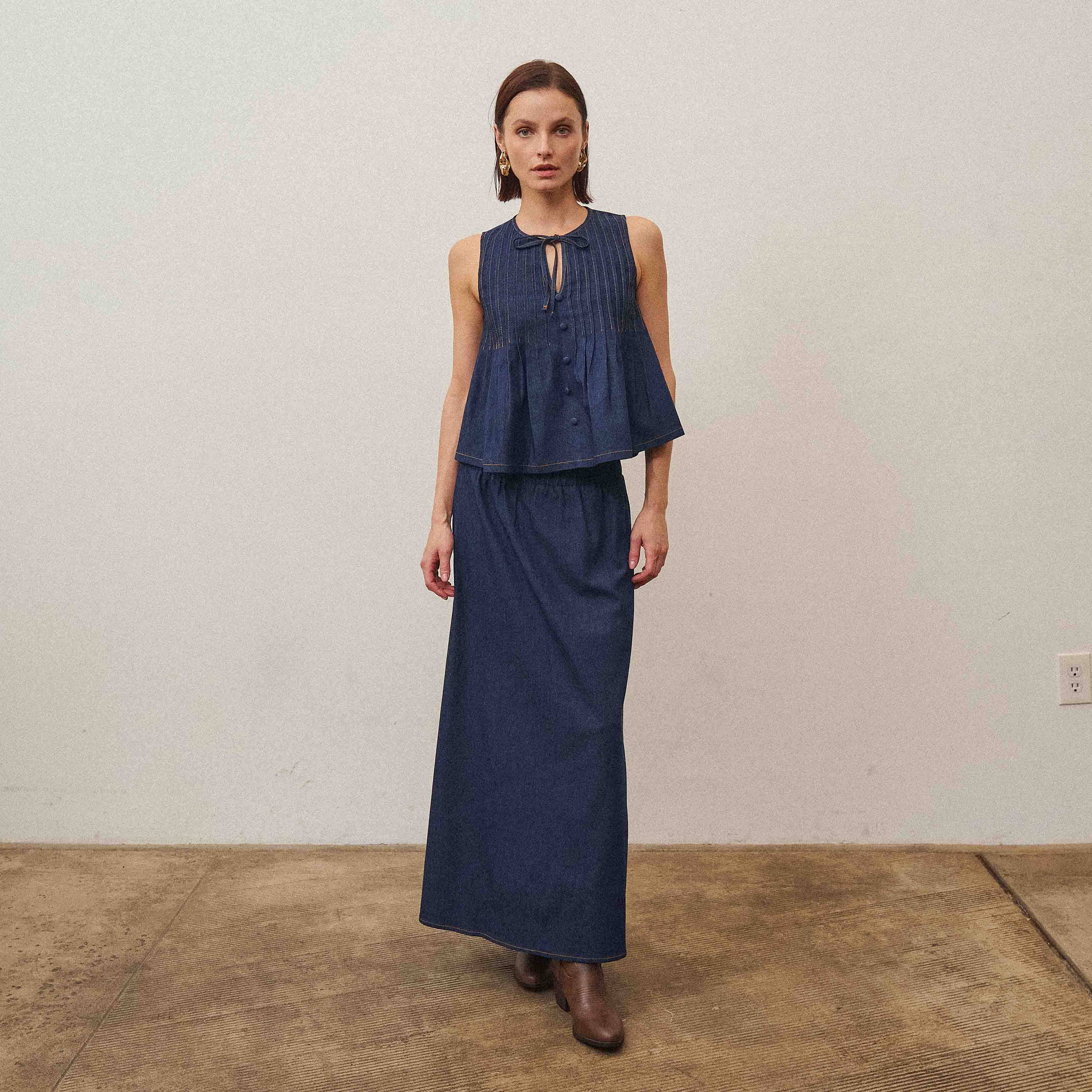 Woman wearing a navy blue sleeveless top and long skirt standing against a plain wall.