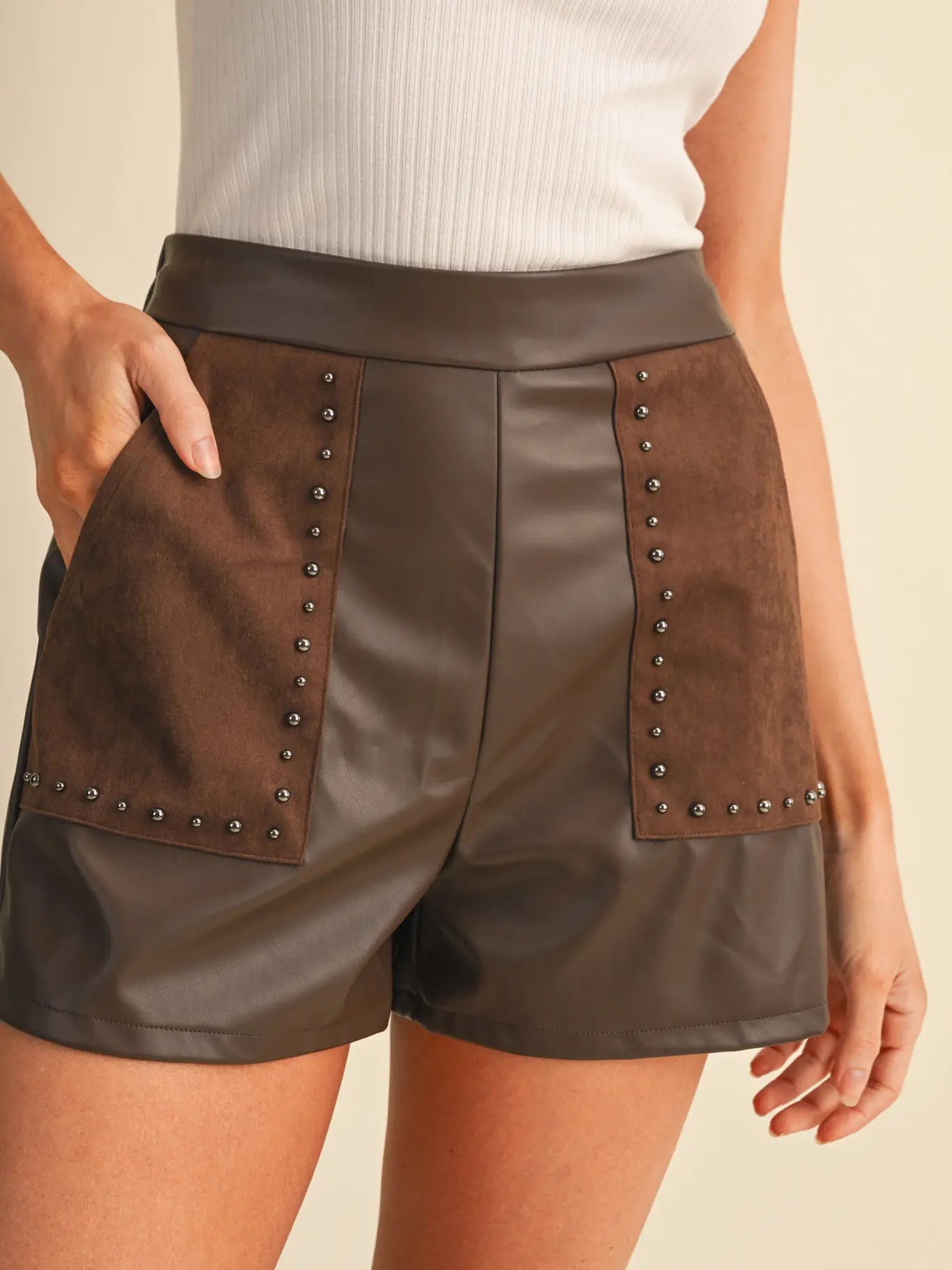 A close-up of a model wearing brown faux-leather shorts with suede-like front patch pockets and rows of silver-tone studs, paired with a white ribbed top against a light beige background.