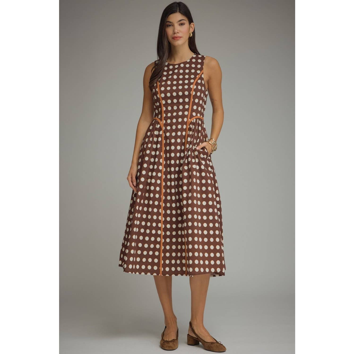 A woman modeling a sleeveless brown and white polka dot midi dress with tan trim, standing against a neutral gray studio background.