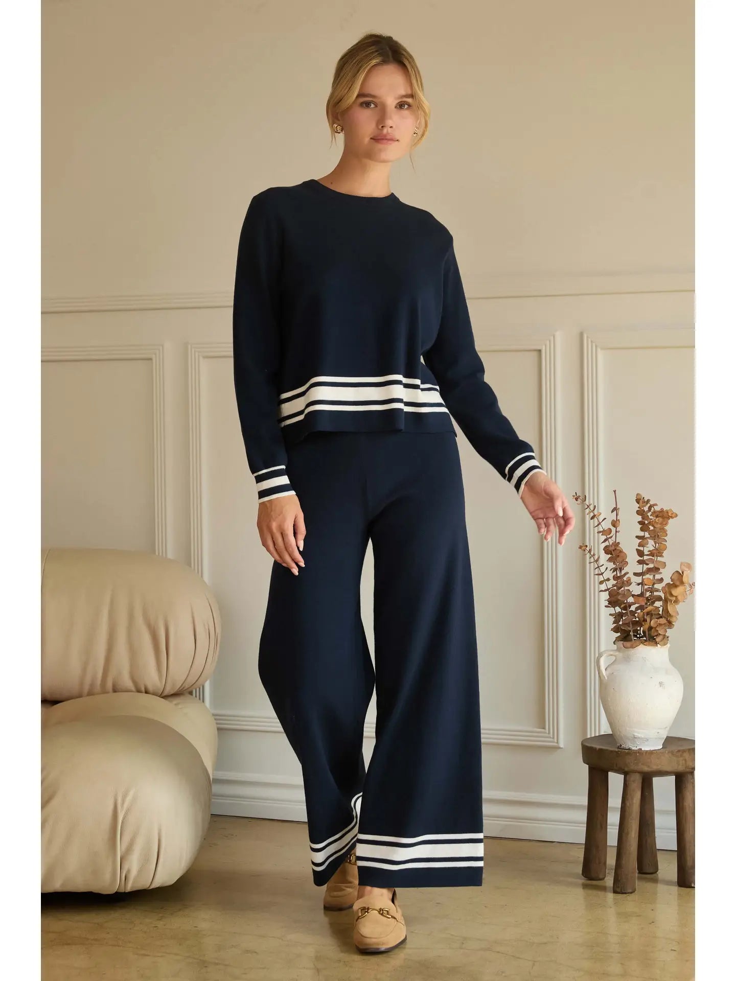 Woman wearing a navy sweater and pants with white stripes in a room with beige walls and wooden flooring.