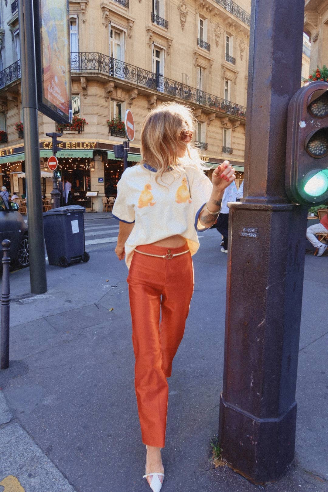 Woman in a white shirt and orange pants standing on a city street with a traffic light.