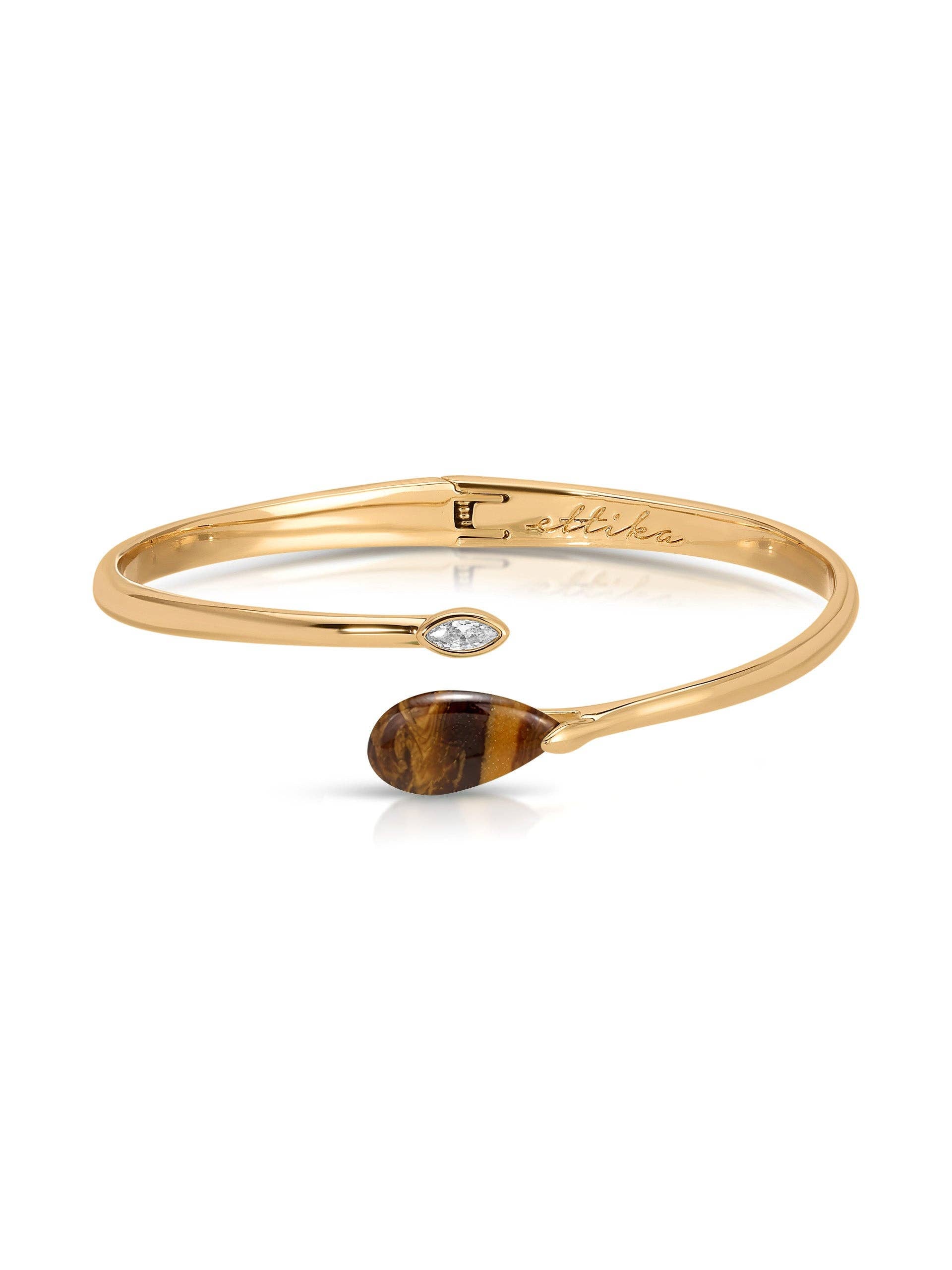 A gold open cuff bangle bracelet featuring a small marquise-cut clear stone on one side and a teardrop-shaped brown tiger's eye stone on the other, displayed on a clean white background.